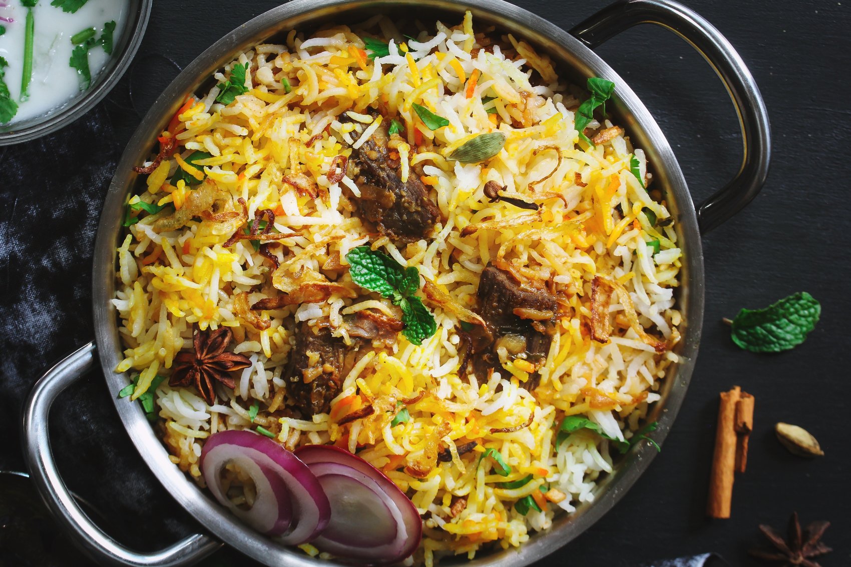 delicious hotel daawat colourful mutton biryani beautifully presented with fried onions, raw onions, star anise, cloves and  in a pan on a black background